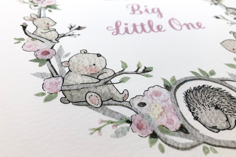 May include: A watercolor illustration of a wreath with a bear, a rabbit, and a hedgehog. The wreath is made of branches and flowers. The text "Big little one" is written in pink lettering.