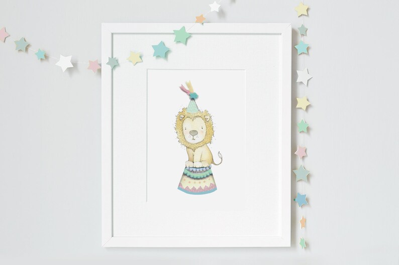 May include: A framed watercolour print of a lion wearing a party hat and sitting on a circus-style platform. The artwork is in a white frame, and pastel star garlands decorate the wall. The lion is light yellow with a light brown mane.