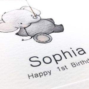 May include: A white card with a black and gray illustration of an elephant holding a string. The text "Sophia Happy 1st Birthday" is printed below the elephant.