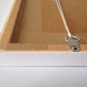 May include: White wooden picture frame with a brown paper backing and a metal hanging hook attached to the back. The hook is secured with a screw.
