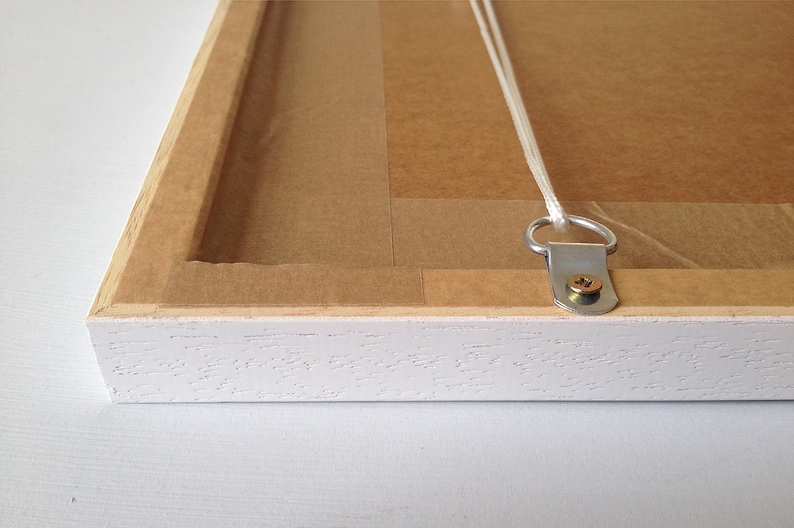 May include: White wooden picture frame with a brown paper backing and a metal hanging hook attached to the back. The hook is secured with a screw.