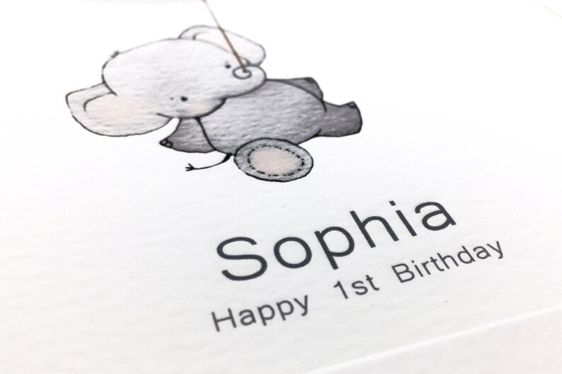 Peut inclure: Une carte blanche avec une illustration noire et grise d'un &eacute;l&eacute;phant tenant une ficelle. Le texte "Sophia Happy 1st Birthday" est imprim&eacute; sous l'&eacute;l&eacute;phant.