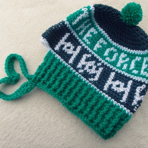May include: Green and blue crocheted hat with a white pom-pom on top. The hat has a green crocheted strap that is tied in a heart shape.