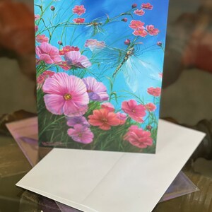 May include: A greeting card featuring a watercolour painting of pink cosmos flowers and two dragonflies against a blue sky background.