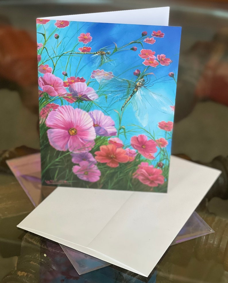 May include: A greeting card featuring a watercolour painting of pink cosmos flowers and two dragonflies against a blue sky background.