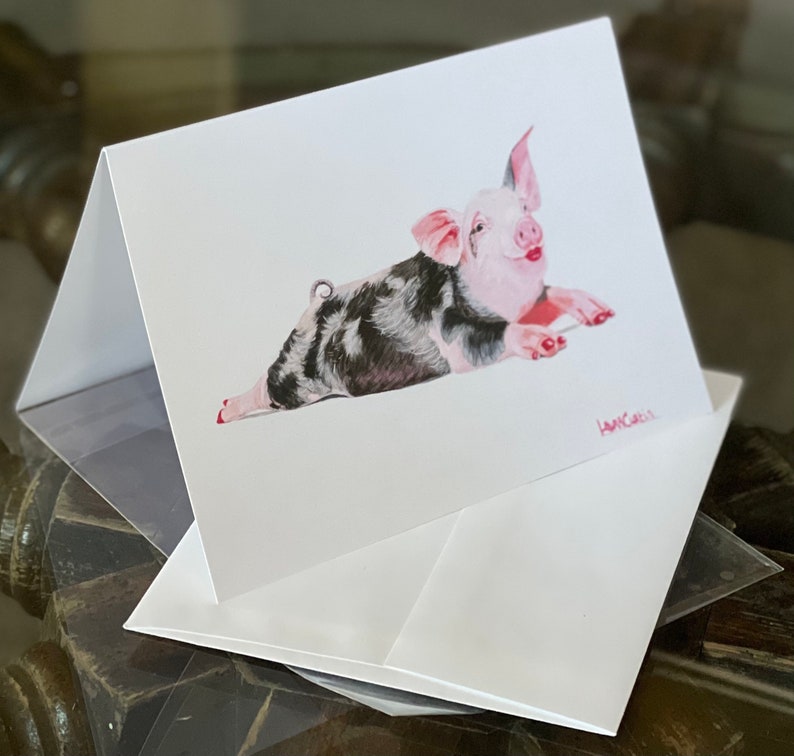 Peut inclure: Une carte blanche avec un cochon noir et blanc couch&eacute; sur le c&ocirc;t&eacute;, les pattes avant tendues. Le cochon a des oreilles roses et un groin rose. La carte est sign&eacute;e "AMY CARL" en bas &agrave; droite.