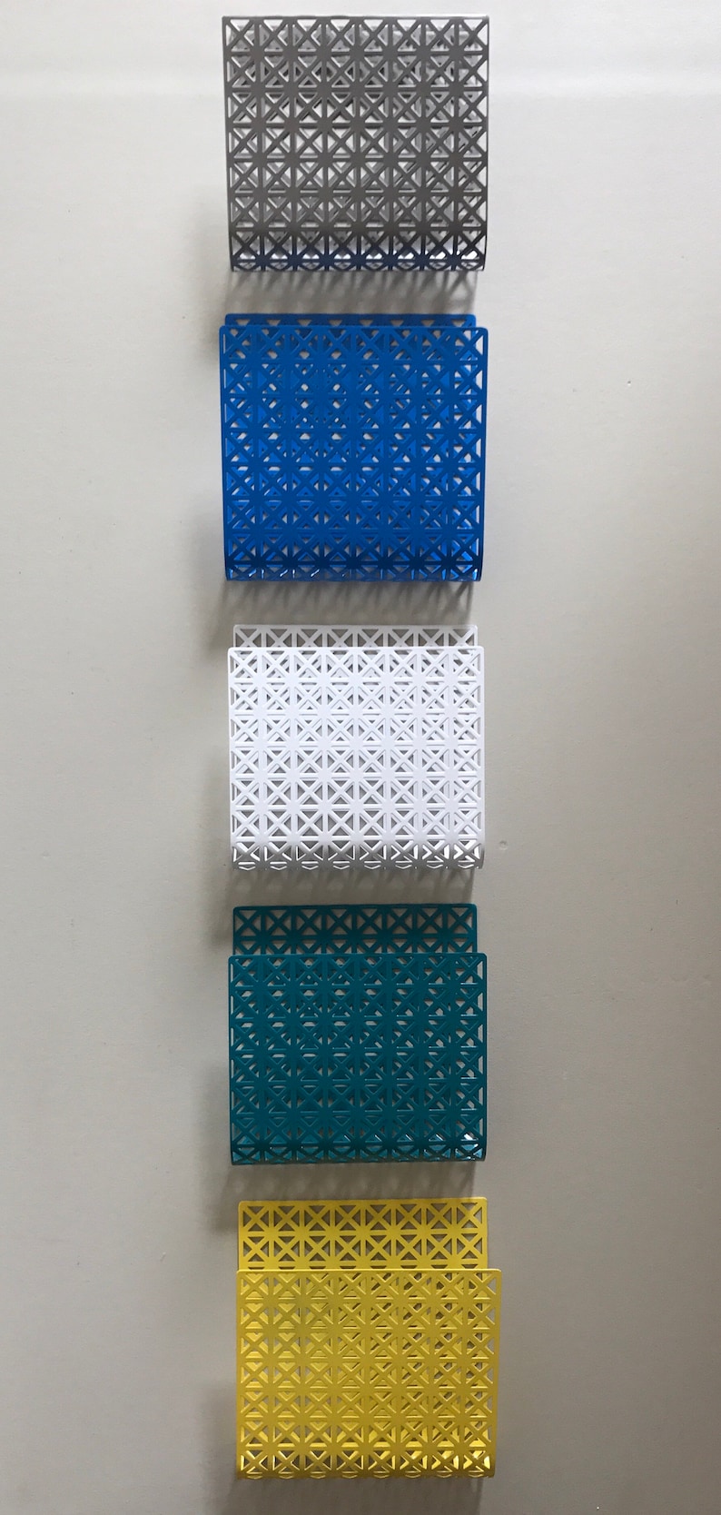 May include: Five metal magazine holders in different colors: gray, blue, white, teal, and yellow. Each holder has a geometric pattern.