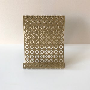 May include: A gold metal bookend with a geometric pattern. The bookend has a stand that is angled to hold books upright.