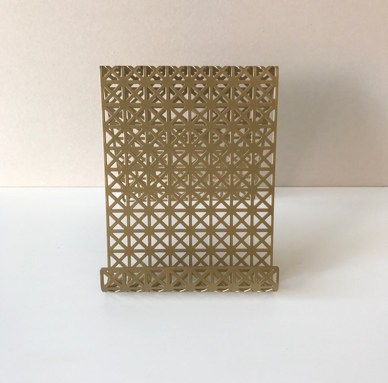 May include: A gold metal bookend with a geometric pattern. The bookend has a stand that is angled to hold books upright.