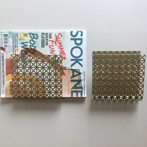 May include: A gold metal magazine rack with a geometric pattern. The rack is holding a magazine with the title "Spokane Summer Fun" on the cover.
