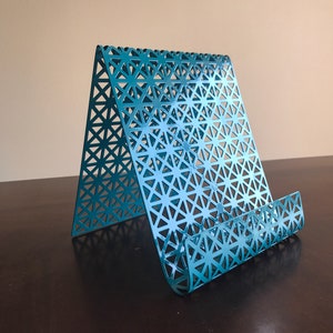 May include: Teal metal phone stand with a geometric pattern. The stand is designed to hold a phone upright.