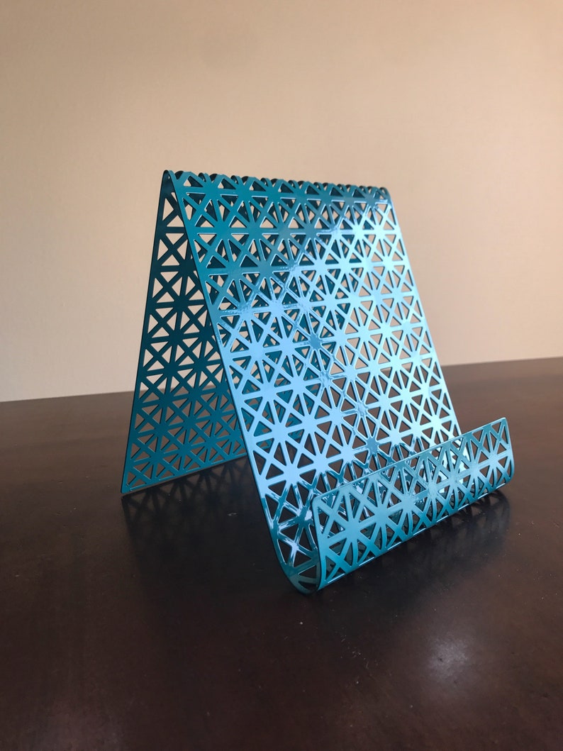 May include: Teal metal phone stand with a geometric pattern. The stand is designed to hold a phone upright.