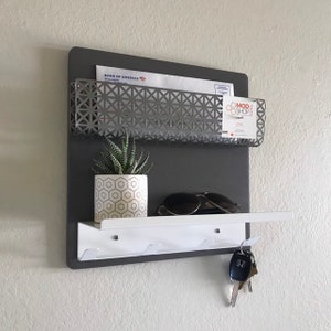Könnte beinhalten: Ein grauer, an der Wand montierter Organizer mit einem Metall-Briefschlitz, einem weißen Regal und Haken für Schlüssel. Der Briefschlitz hat ein geometrisches Muster und das Regal einen weißen Hintergrund. Auf dem Regal steht eine kleine Topfpflanze und eine Sonnenbrille. An den Haken hängen Schlüssel. Der Text "MOD SHOP" ist auf dem Briefschlitz sichtbar.