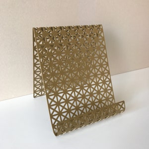 May include: A gold metal phone stand with a geometric pattern. The stand is designed to hold a phone or tablet upright.