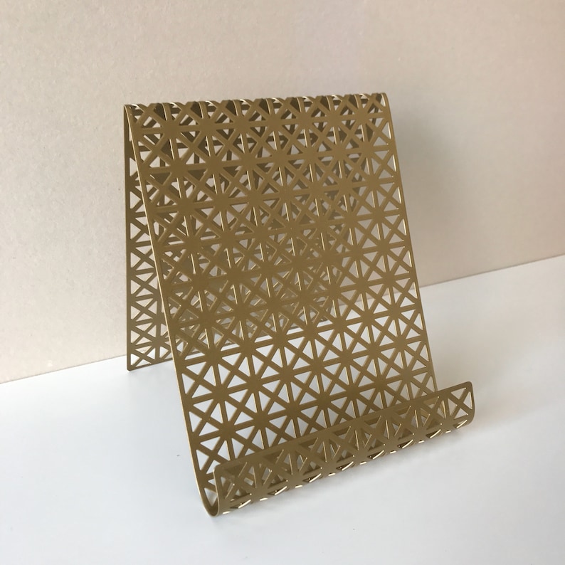 May include: A gold metal phone stand with a geometric pattern. The stand is designed to hold a phone or tablet upright.