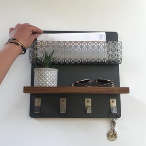 May include: A wall-mounted organizer with a dark gray backing, a metal mail basket, a wooden shelf, and four hooks. A small potted plant, sunglasses, and keys hang from the organizer. The key chain has a camper design.