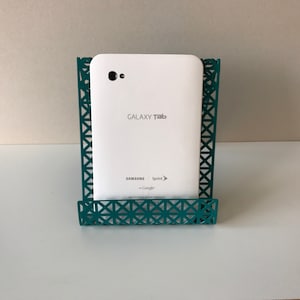 May include: White Samsung Galaxy Tab tablet in a teal metal stand. The tablet has the text "GALAXY Tab" and "SAMSUNG" on the back. The stand has a geometric pattern.