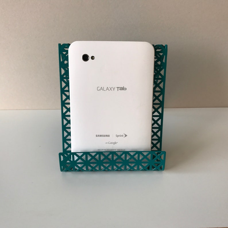 May include: White Samsung Galaxy Tab tablet in a teal metal stand. The tablet has the text "GALAXY Tab" and "SAMSUNG" on the back. The stand has a geometric pattern.