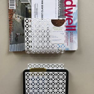 May include: Two white metal wall organizers with a geometric pattern. The top organizer holds a magazine and a business card. The bottom organizer holds a black tablet.