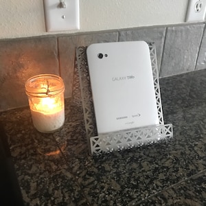 May include: A white Samsung Galaxy Tab tablet is propped up in a silver metal stand. The tablet is displaying the text "GALAXY Tab" and "SAMSUNG" with the Sprint logo and "on Google" text below. The tablet is on a gray countertop with a lit candle in a glass jar to the left.