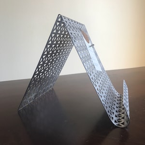 May include: A gray metal stand with a geometric pattern. The stand is designed to hold a book or tablet at an angle.