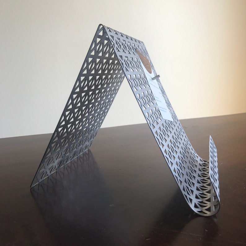 May include: A gray metal stand with a geometric pattern. The stand is designed to hold a book or tablet at an angle.