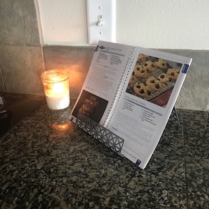 May include: A cookbook with a recipe for chocolate chip cookies is propped open on a metal cookbook stand. The cookbook is on a granite countertop with a lit candle in a glass jar.