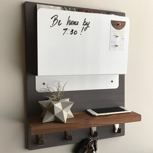 May include: A wall-mounted organizer with a white dry-erase board, a wooden shelf, and metal hooks. The board has the handwritten text "Be home by 7:30!". A geometric planter with a small plant, a smartphone, and keys are on the shelf.