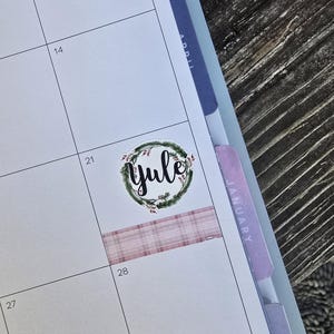 May include: A planner page with a "Yule" sticker surrounded by a green wreath. A pink plaid washi tape strip is placed below the sticker. The calendar is open to January, with the 28th visible.