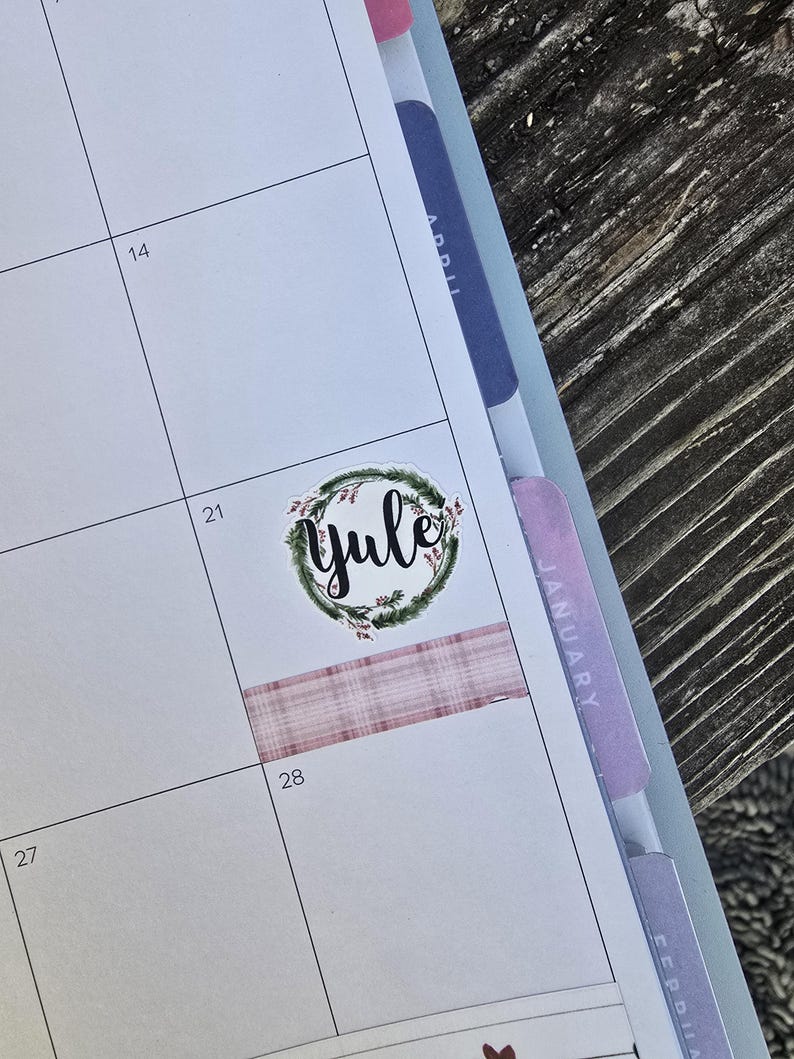 May include: A planner page with a "Yule" sticker surrounded by a green wreath. A pink plaid washi tape strip is placed below the sticker. The calendar is open to January, with the 28th visible.