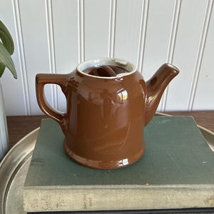 May include: A glossy brown ceramic teapot with a matching lid, a curved handle, and a short spout. The teapot features a white interior rim and is positioned on top of two stacked hardcover books, which are resting on a round silver metal tray.