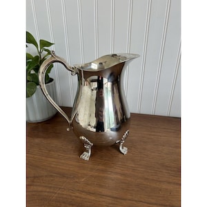 May include: A polished silver-toned metal pitcher featuring a curved handle, a wide pouring spout, and three decorative feet shaped like animal paws. The reflective surface of the pitcher shows a distorted view of the surrounding environment.