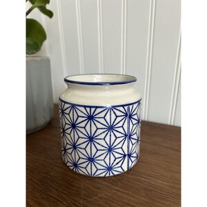 May include: A cylindrical ceramic jar with a cream-colored base and a repeating blue geometric star pattern. The rim is finished with a thin blue line, and the vessel features a smooth, glossy glaze throughout its exterior surface.