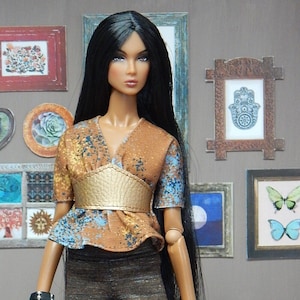 May include: A fashion doll with long black hair wearing a brown and blue patterned blouse with a gold belt, brown and grey striped trousers, and black shoes. The doll is standing in front of a wall with several framed pictures.