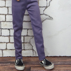 May include: A doll wearing a gray mock neck shirt and purple pants. The doll is also wearing black and white sneakers. The doll is standing in front of a white brick wall.