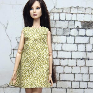Puede incluir: Muñeca de moda con un vestido verde oliva con estampado de lunares blancos. La muñeca tiene pelo largo y oscuro y zapatos verdes a juego. El fondo presenta una pared de ladrillos blancos desgastados y un suelo de madera.