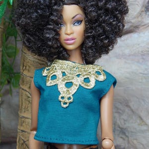 May include: A fashion doll with dark curly hair, wearing a teal top with gold embroidered collar and patterned orange and black pants. The doll has a tan complexion and is posed in front of a neutral background.