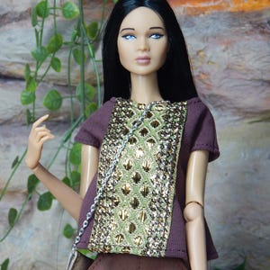 May include: A fashion doll with long black hair and blue eyes wearing a dark brown short-sleeved top with a central panel of green and gold metallic textured fabric, paired with matching dark brown cuffed shorts and a small silver-chained shoulder bag.