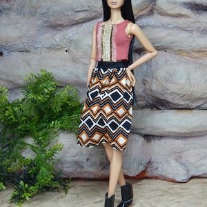 May include: A fashion doll with long black hair wearing a sleeveless mauve crop top with a central gold sequined panel and a black waistband. The doll wears a knee-length skirt featuring a geometric diamond pattern in brown, white, and black, paired with black ankle boots.