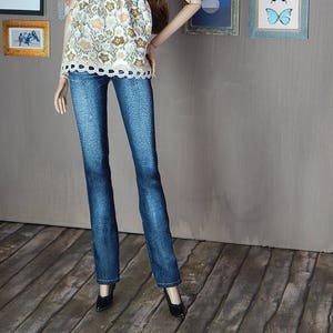 May include: A doll wearing a white and brown floral print tank top with a lace trim, blue jeans, and black heels. The doll has long brown hair and is standing in front of a wall with framed pictures.