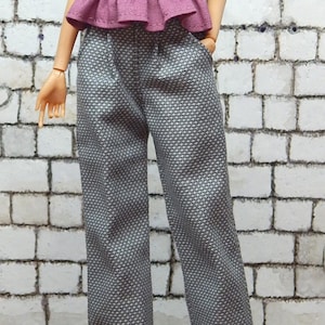 May include: A doll wearing a pink ruffled top with a black belt and grey patterned trousers.
