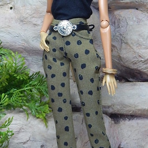 May include: A fashion doll with dark curly hair wearing a black short-sleeved top and olive green trousers featuring a black polka dot pattern. The doll is accessorised with a black belt featuring a silver circular buckle and black high-heeled shoes.