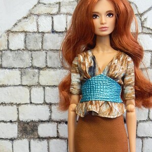May include: A doll with long red hair wearing a brown skirt, a brown and white patterned blouse, and a blue belt. The doll is standing in front of a white brick wall.