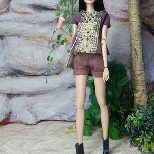 May include: A fashion doll with long black hair wearing a brown short-sleeved top with a central gold sequined panel, matching brown shorts, and black ankle boots. The doll carries a small brown shoulder bag with a silver chain strap.