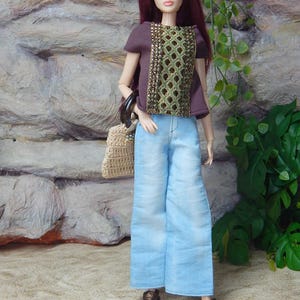 May include: A fashion doll with long burgundy hair wearing a dark brown short-sleeved top with a central gold and green patterned panel, light blue wide-leg denim pants, and metallic bronze shoes, while carrying a small beige woven handbag.
