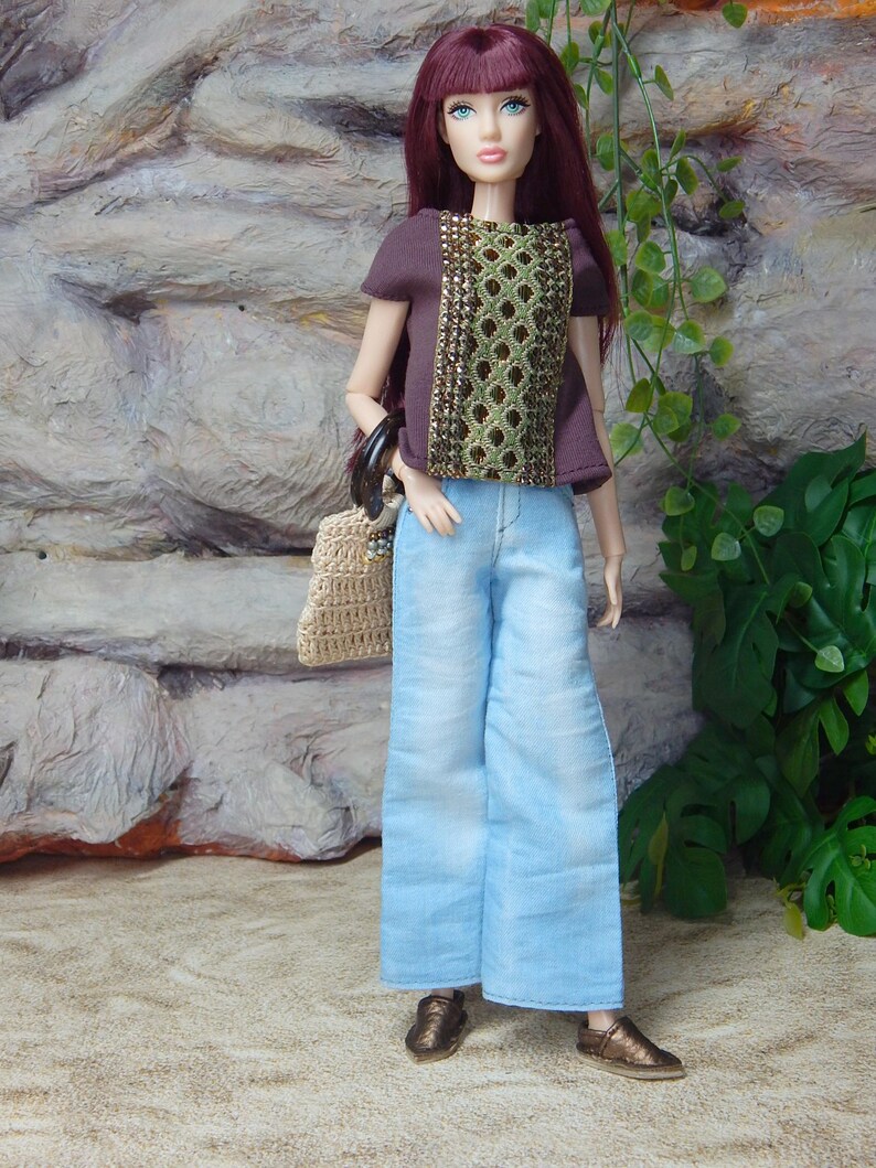 May include: A fashion doll with long burgundy hair wearing a dark brown short-sleeved top with a central gold and green patterned panel, light blue wide-leg denim pants, and metallic bronze shoes, while carrying a small beige woven handbag.