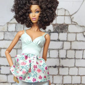 May include: A doll with brown skin and a large afro hairstyle is wearing a light blue and white floral print top with a light blue denim skirt.