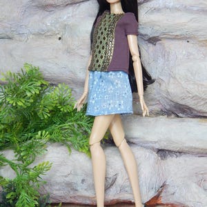 May include: A fashion doll with long dark hair wearing a brown short-sleeved top with a central green and gold diamond-patterned panel, a light blue skirt featuring a white floral print, and black high-heeled shoes with pointed toes.