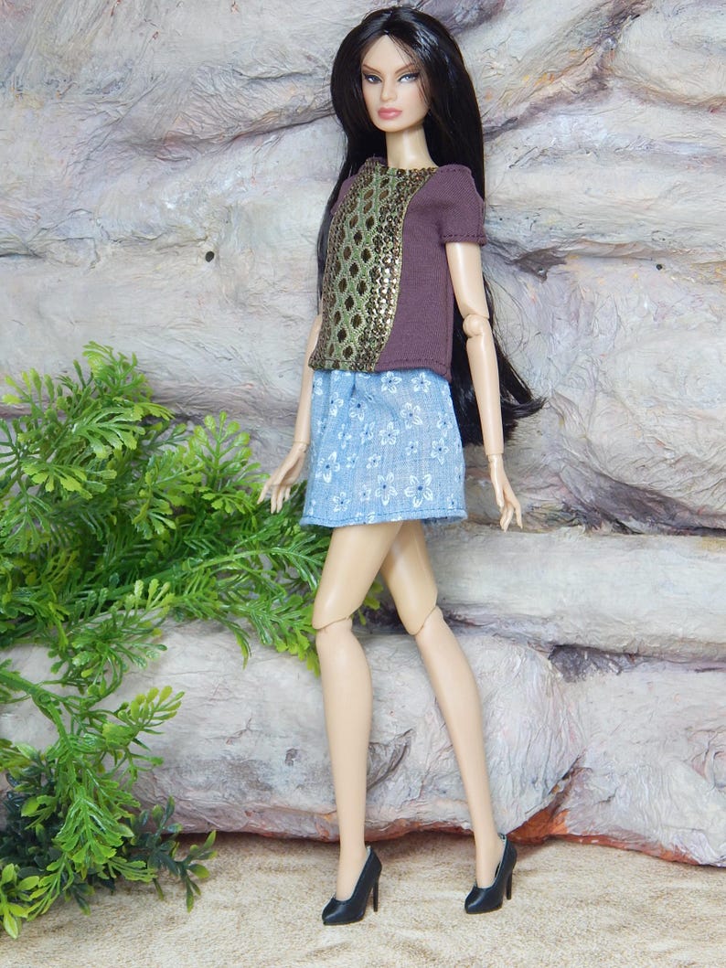 May include: A fashion doll with long dark hair wearing a brown short-sleeved top with a central green and gold diamond-patterned panel, a light blue skirt featuring a white floral print, and black high-heeled shoes with pointed toes.