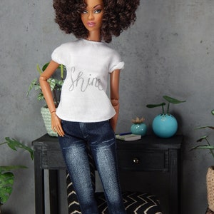 May include: A doll with brown curly hair wearing a white t-shirt with the word "shine" printed on it and blue jeans.